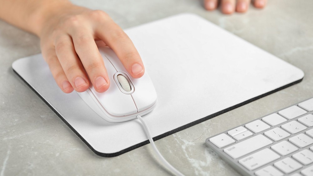 Woman uses a wired computer mouse on a white mouse pad at a light grey marble table.